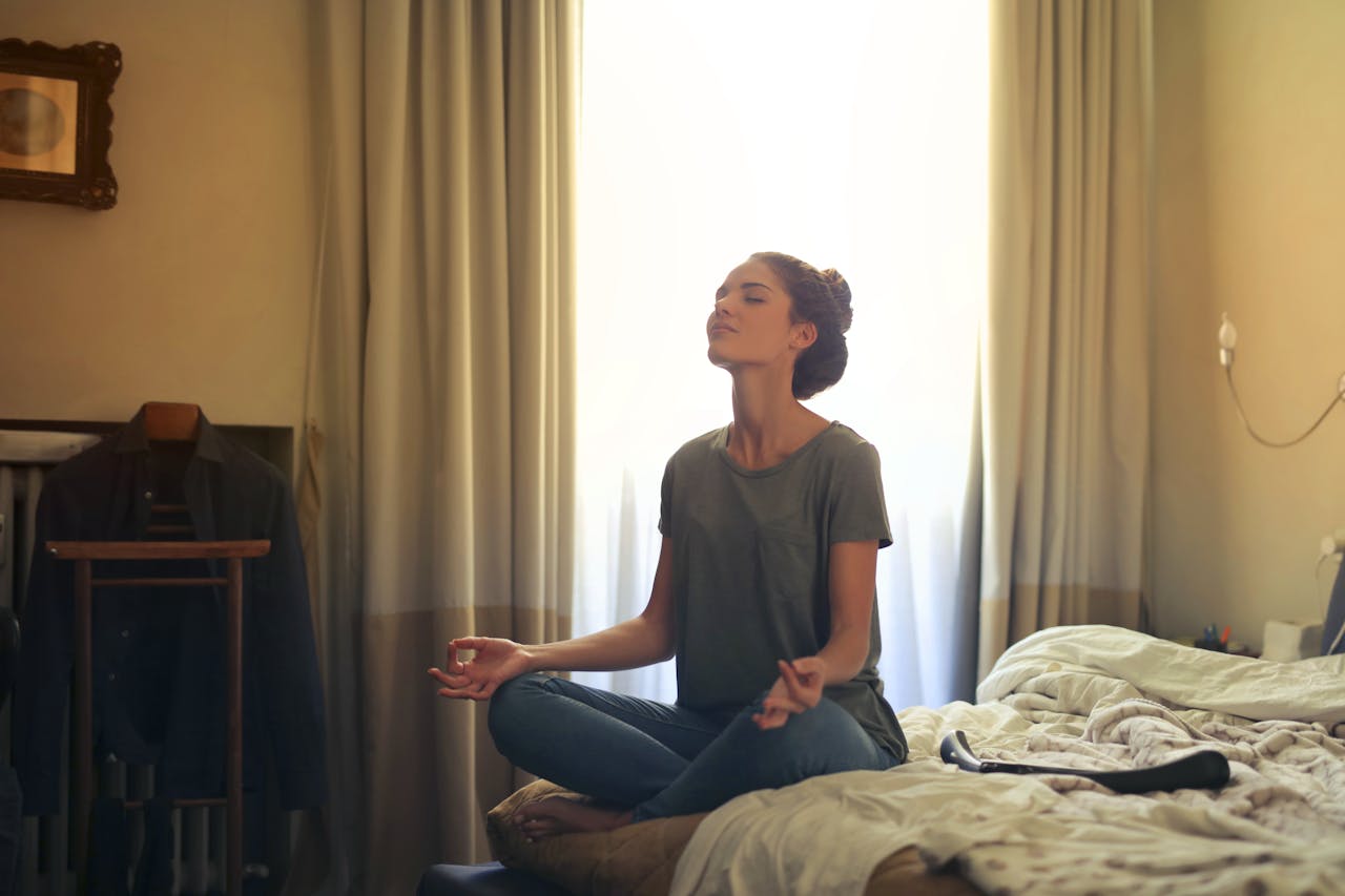 about-us Adult woman practicing meditation on her bed surrounded by a calm bedroom atmosphere.